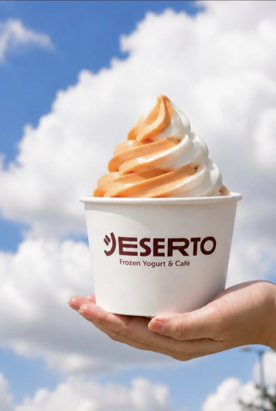 An orange-and-vanilla creamsicle frozen yogurt swirl in a Deserto cup, held up against a bright blue cloudy sky