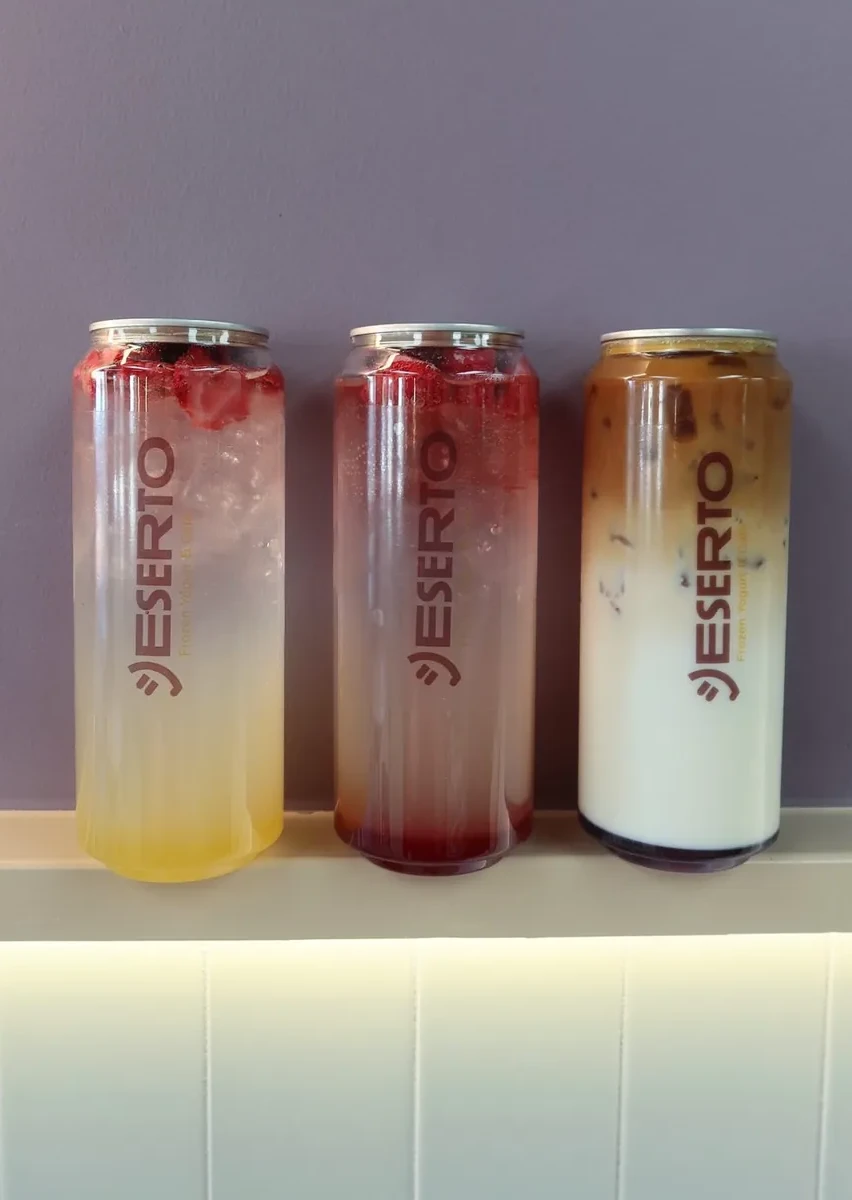 Three Deserto sparkling fruit tonics in branded cans lined up on a lit shelf against the café's lavender wall