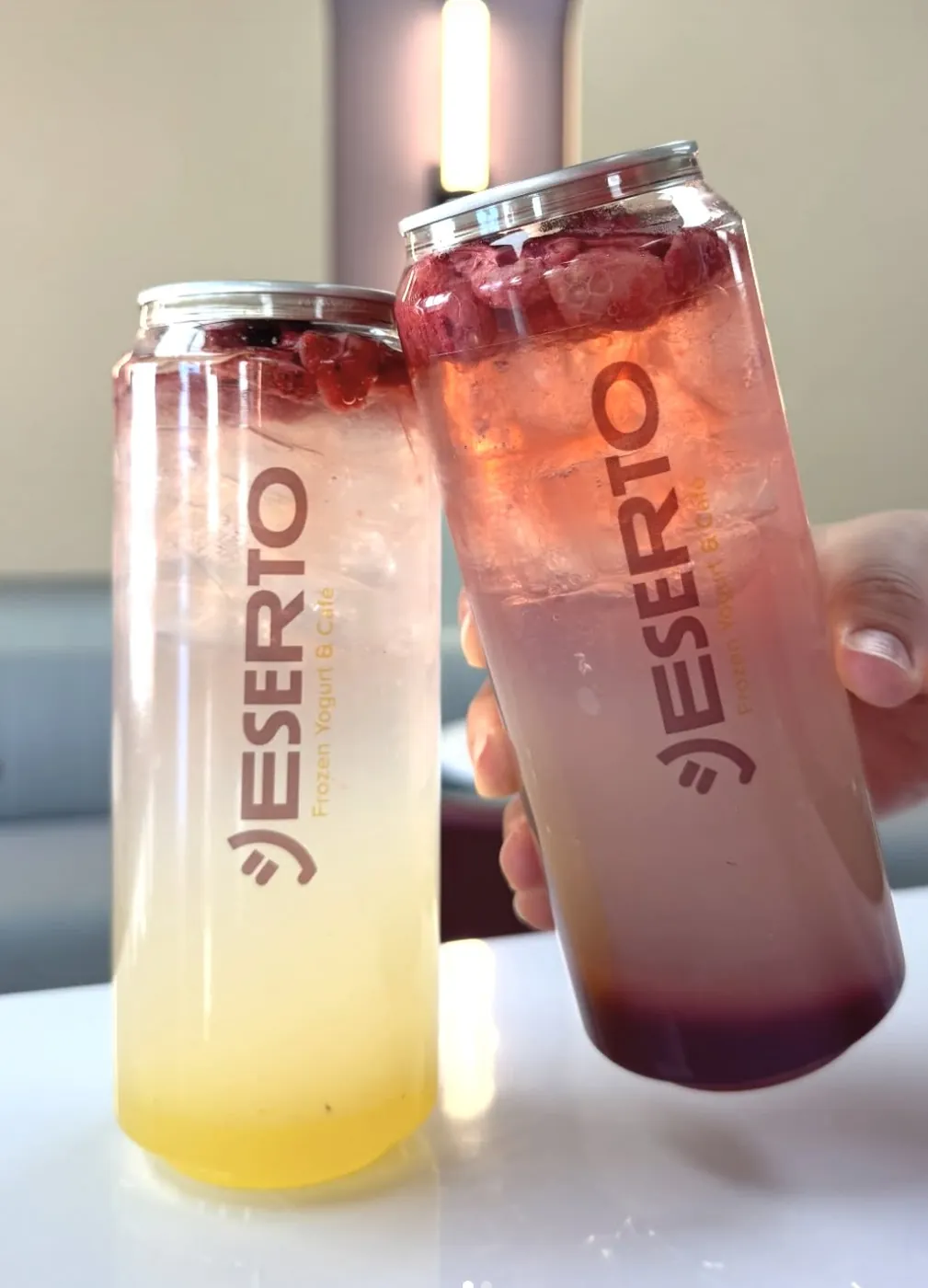 Two friends toasting with berry Deserto tonics in branded cans inside the warm, sconce-lit café